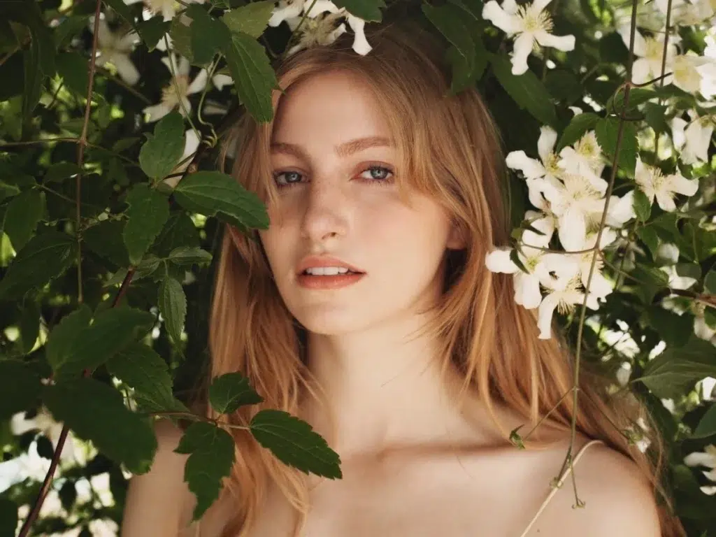 Young woman with long blonde hair among green leaves and white blossoms, facing the camera with a soft expression